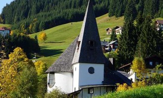 A picturesque church with a tall spire stands in a green landscape. In the background, there are gentle hills and some houses. | © Ev. Kirche Kleinwalsertal