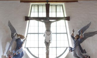 A quiet altar in a chapel with a cross and sculptures of angels. The windows let in a lot of light and create a peaceful atmosphere. | © Ev. Kirche Kleinwalsertal