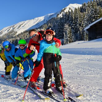 A group of children and an adult skiing in the snowy mountain landscape. They are wearing colorful ski clothes and smiling at the camera. | © Andy Herr | Werbewind GmbH