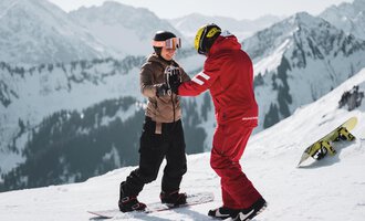 Zwei Snowboarder stehen im Schnee und helfen sich gegenseitig beim Lernen. Im Hintergrund sind schneebedeckte Berge zu sehen.